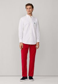 Hombre con una camisa blanca de botones con un pequeño logo, pantalones rojos y zapatillas negras, de pie contra un fondo gris claro liso.