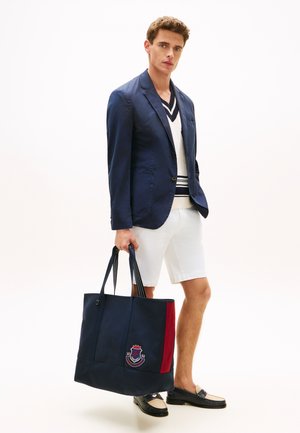 Homme portant un blazer bleu marine, un short blanc et des mocassins, tenant un grand sac cabas bleu marine avec une bande rouge et un emblème de blason.