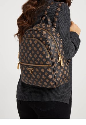 Femme portant un pull gris foncé et un pantalon noir, portant un sac à dos de marque noir et marron avec des fermetures éclair dorées et des accents de logo.