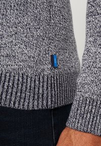 Pull en maille dans des tons gris avec un ourlet côtelé. Présente une étiquette de marque bleue. Gros plan sur la texture de la manche et de l'ourlet.