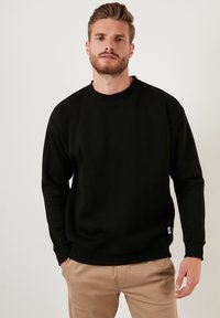 Sweatshirt negru din bumbac, cu mâneci lungi, guler rotund, manșete și tiv din jerse. Purtați peste pantaloni bej, cu un mic patch cu logo în partea de jos.