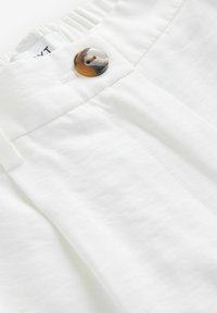 Gros plan sur la ceinture d'un pantalon blanc avec un bouton rond marron et un détail de couture visible.