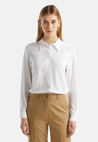 Bílá blůza s dlouhým rukávem a zapínáním na knoflíky vpředu, s klasickým límcem, vyrobená z hladkého materiálu. Spárována s béžovými kalhotami.