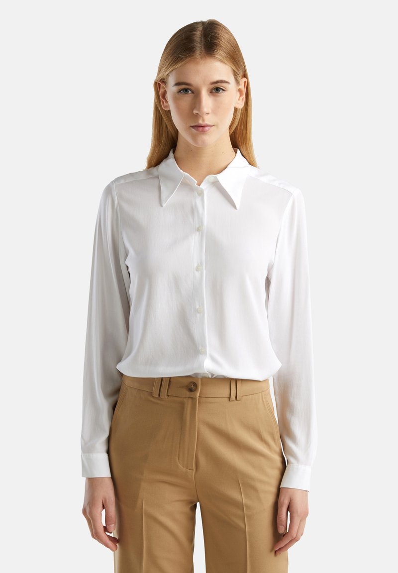 Bílá blůza s dlouhým rukávem a zapínáním na knoflíky vpředu, s klasickým límcem, vyrobená z hladkého materiálu. Spárována s béžovými kalhotami.
