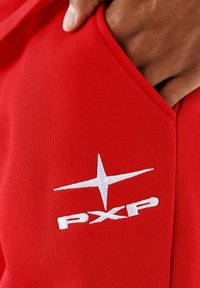 Hand glijdt in de zak van een rode stof met een witte geborduurde PXP-logo en ster symbool.