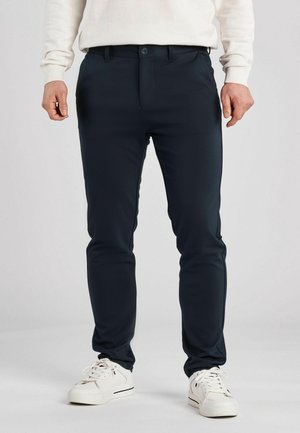 Man die slim fit donkerblauwe broek, witte sneakers en een wit shirt met lange mouwen draagt, staand tegen een effen lichte achtergrond.