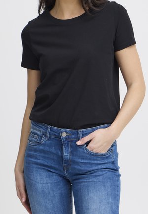 Kobieta ubrana w czarny t-shirt z krótkim rękawem wpuszczony w niebieskie jeansy, z jedną ręką w przedniej kieszeni, a drugą przy boku.