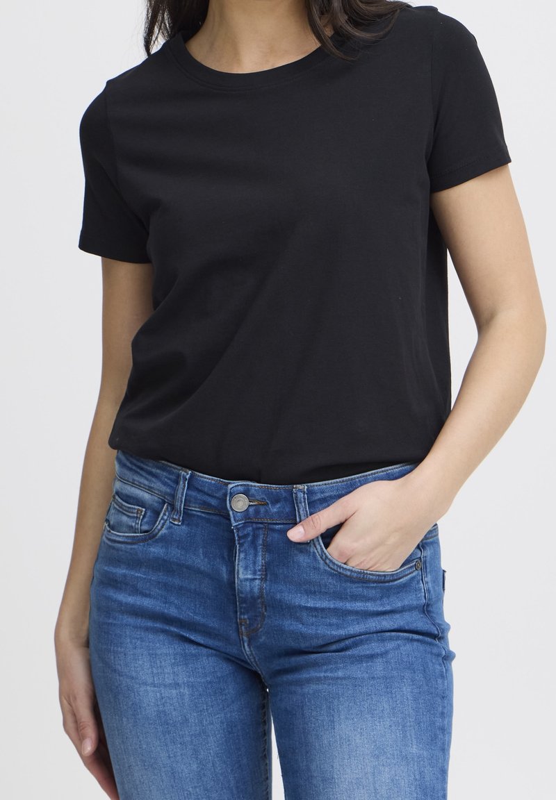 Femme portant un t-shirt noir à manches courtes rentré dans un jean bleu, une main dans la poche avant et l'autre le long du corps.