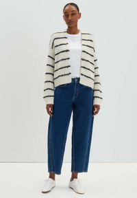 Strickjacke in Creme mit schwarzen Streifen, kürzedes Design, grobe Textur. Kombiniert mit blauen High-Waist-Jeans und weißen Sneakern.