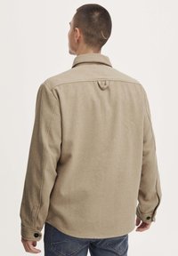 Beige Wollhemdjacke mit auffälligem Kragen, geknöpften Manschetten und einer Schlaufenverzierung auf der Rückseite; kombiniert mit dunklen Jeans.
