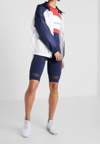 Giacca a vento sportiva bianca e blu navy, abbinata a pantaloni bike blu navy con pannelli in rete. Le scarpe sono di design in rete bianca.
