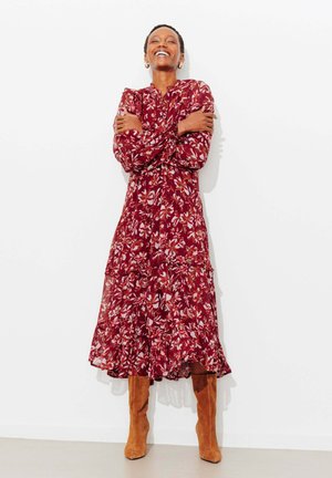 Femme souriante en robe longue rouge à fleurs et bottes en daim marron clair, debout les bras croisés contre un mur blanc.