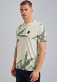 T-shirt beige clair avec un imprimé de feuilles vert foncé, col rond, manches courtes et un petit patch logo noir sur la poitrine.