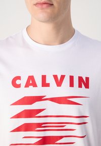 Biely bavlnený tričko s výrazným červeným textom "CALVIN" nad abstraktnými červenými tvarmi, mierne voľný strih, s dizajnom okrúhleho výstrihu.