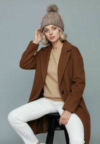 Brown woolen coat over a beige sweatshirt, white jeans, and a tan knitted beanie with a pom-pom. Subject sitting on a black stool.