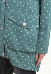 Wodoodporna kurtka w kolorze zielonkawoniebieskim z jasnoniebieskimi kropkami, z zapięciem na guziki, kieszenią na piersi i kontrastową fakturą materiału.