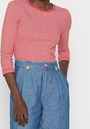 Person trägt ein rot-weiß gestreiftes Langarmoberteil, das in eine hoch taillierte blaue Hose mit Knöpfen und Falten gesteckt ist.