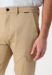 Khaki cargobroek met een gladde stoftextuur, een knoopsluiting en een zijzak. Calvin Klein branding zichtbaar bij de tailleband.