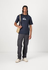 Mørkeblå t-skjorte med grå logo, kombinert med grå cargo-bukser med sidelommer og en beige skulderveske. Lysfarget fottøy.