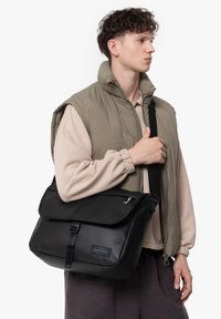 Svart budväska med ett lock, tillverkad av texturerad nylon, med en sidoberlock och justerbar rem, hålls av en person iförd en vadderad väst.