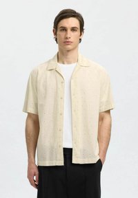 Camicia a maniche corte con bottoni realizzata in un tessuto leggero e testurizzato di un beige chiaro, con piccoli pattern a occhiello, abbinata a una t-shirt bianca.