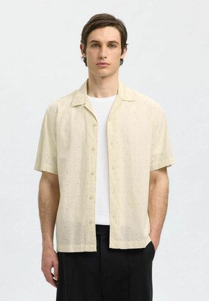 Camicia a maniche corte con bottoni realizzata in un tessuto leggero e testurizzato di un beige chiaro, con piccoli pattern a occhiello, abbinata a una t-shirt bianca.