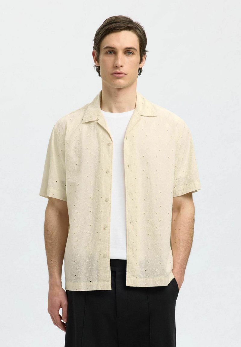 Camicia a maniche corte con bottoni realizzata in un tessuto leggero e testurizzato di un beige chiaro, con piccoli pattern a occhiello, abbinata a una t-shirt bianca.