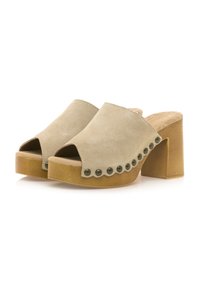Mules beige de ante abiertos con un tacón de madera grueso. Detalle de borde ondulado con tachuelas decorativas en contraste a lo largo de un lado.