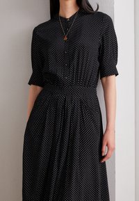 Kobieta w czarnej sukience z krótkim rękawem, zapinanej na guziki, w białe kropki, oraz złotym naszyjniku z okrągłym, czerwonym wisiorkiem.