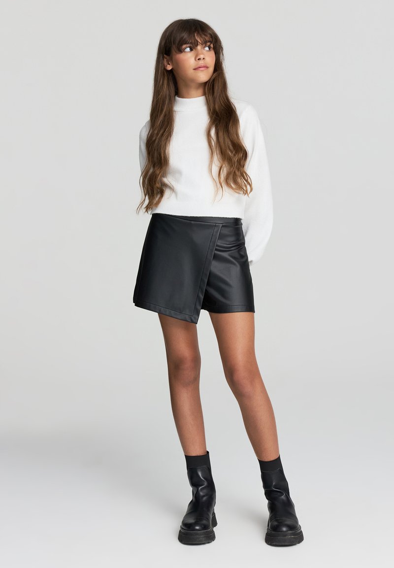 Mini gonna asimmetrica nera in finta pelle, texture liscia, abbinata a un maglione bianco a maniche lunghe e stivali neri alla caviglia.