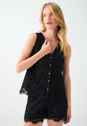 Vrouw met blond haar die een zwart mouwloos kanten topje en bijpassende kanten shorts draagt, kijkt naar links.
