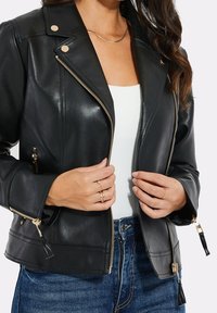 Veste de moto en cuir noir avec des accents en métal doré, présentant un design ajusté, des poches zippées et des franges décoratives sur les fermetures éclair.