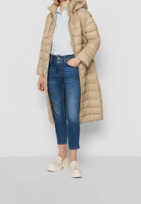 Béžový dlouhý zateplený kabát s kapucí, prošívaný design, kombinovaný s modrými zkrácenými dž�íny a béžovými platformovými mokasíny s třásněmi.