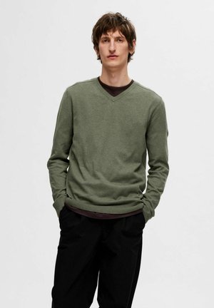 Grøn langærmet V-hals sweater lavet af blødt stof, med ribbede manchetter og kant, lagdelt over en mørk skjorte. Sorte bukser fuldender looket.