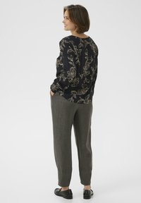 Donna che indossa una blusa nera a fiori, pantaloni grigi e mocassini neri, in piedi con le mani in tasca e girata di lato su uno sfondo bianco.