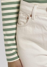 Pantalon en denim beige avec une poche avant et des accessoires en cuivre, associé à un haut ajusté à rayures vertes et crème à manches longues.