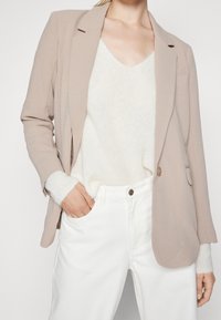 Světle béžový blejzr s klopami, na jedno knoflíkové zapínání, nošený přes bílý svetr a bílé džíny, ukazuje uvolněný střih a jemnou texturu.