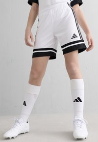 Persona vestida con pantalones cortos de fútbol Adidas en blanco y negro, calcetas altas blancas de Adidas y zapatillas blancas, posando frente a un fondo simple.