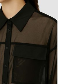 Camicetta nera trasparente con colletto classico, chiusura a bottoni e taschino sul petto con bordo nero solido e base in tessuto testurizzato.