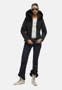 Giacca nera con cappuccio in pelliccia sintetica, abbinata a una maglietta bianca e jeans scuri. Il modello indossa occhiali da sole e stivali chiari con lacci.