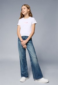 Vit kortärmad t-shirt med en subtil rosa grafik, kombinerad med vidbenade blå denimjeans och vita sneakers. Slät textur, avslappnad design.