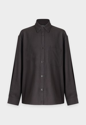 Uomo in piedi che indossa una camicia scura larga con bottoni, pantaloni neri a gamba larga e scarpe nere su uno sfondo chiaro semplice.