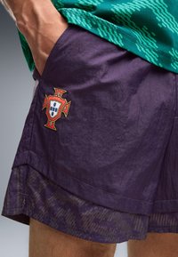 Homme portant un short de sport violet avec le blason du football du Portugal et une chemise verte à motifs, main dans la poche.