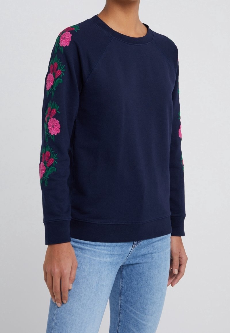Sweat-shirt bleu marine avec broderie florale rose sur les deux manches, porté avec un jean bleu clair sur une personne debout.
