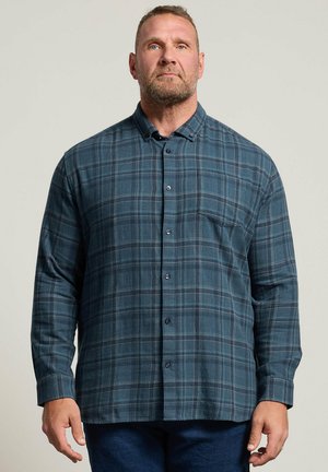 Homme avec une barbe portant une chemise à carreaux bleue boutonnée, debout devant un fond clair uni.