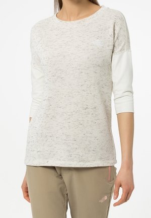 Femme portant un haut à manches longues gris clair en tissu texturé et un pantalon beige avec un petit logo sur la cuisse.