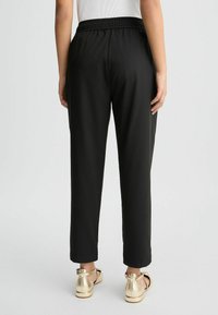Schwarze, locker sitzende Hosen mit elastischem Bund, glatter Textur und tailliertem Design; getragen mit sandalen mit goldenen Akzenten.