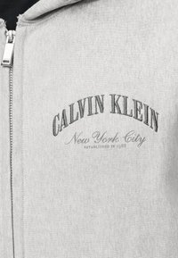 Svijetlosiva majica s kapuljačom na patentni zatvarač s velikim slovima "CALVIN KLEIN" i izvezenim natpisima "New York City" te "Established in 1968" na prsima.