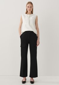 Weißes ärmelloses Oberteil mit einem gefältelten Ausschnitt, kombiniert mit schwarzen Cargo-Hosen mit Seitentaschen und einem taillierten Bund. Schwarze Ballerinas mit quadratischer Zehenform.