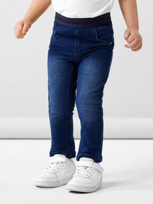 Pantalones de mezclilla azul oscuro con una textura suave, con una cinturilla elástica negra brillante y un diseño clásico de cinco bolsillos, combinados con zapatillas blancas.